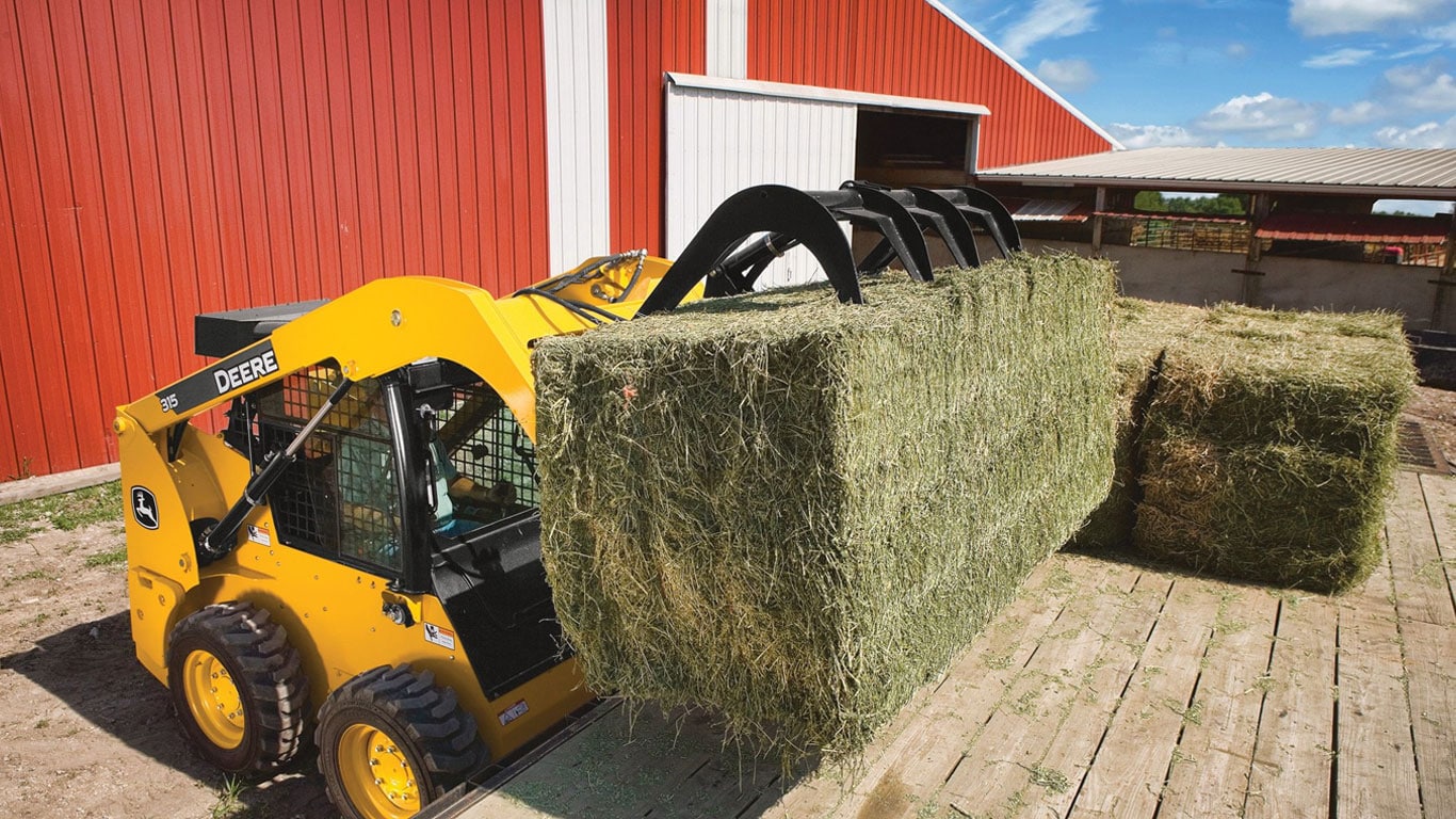 Une chargeuse à direction différentielle John Deere équipée de l’accessoire pinces pour rebuts transporte de la paille.